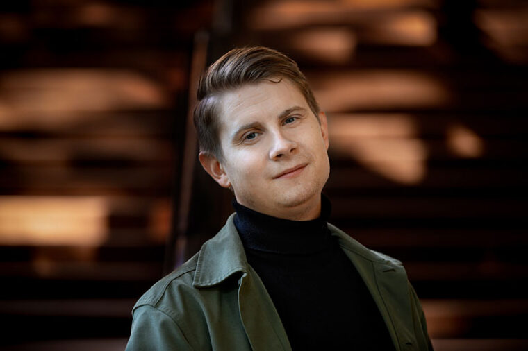 Foto av regissör David Sandqvist. Porträtt av en person med kort, bakåtkammat hår, klädd i svart polotröja och grön jacka, stående framför en mjukt suddig bakgrund med varma ljusformer.