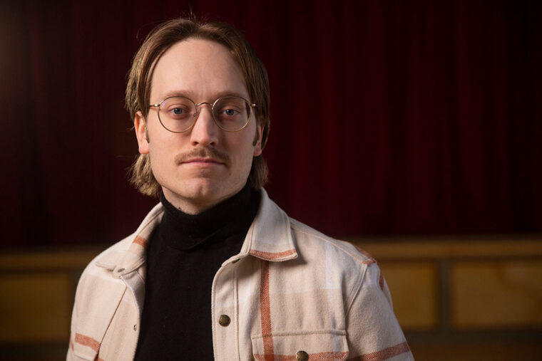 Fotografi av författaren Axel Åhman. Porträtt av en person med runda glasögon och mustasch, klädd i svart polotröja och ljus rutig jacka, stående framför en mörkröd sammetsbakgrund.